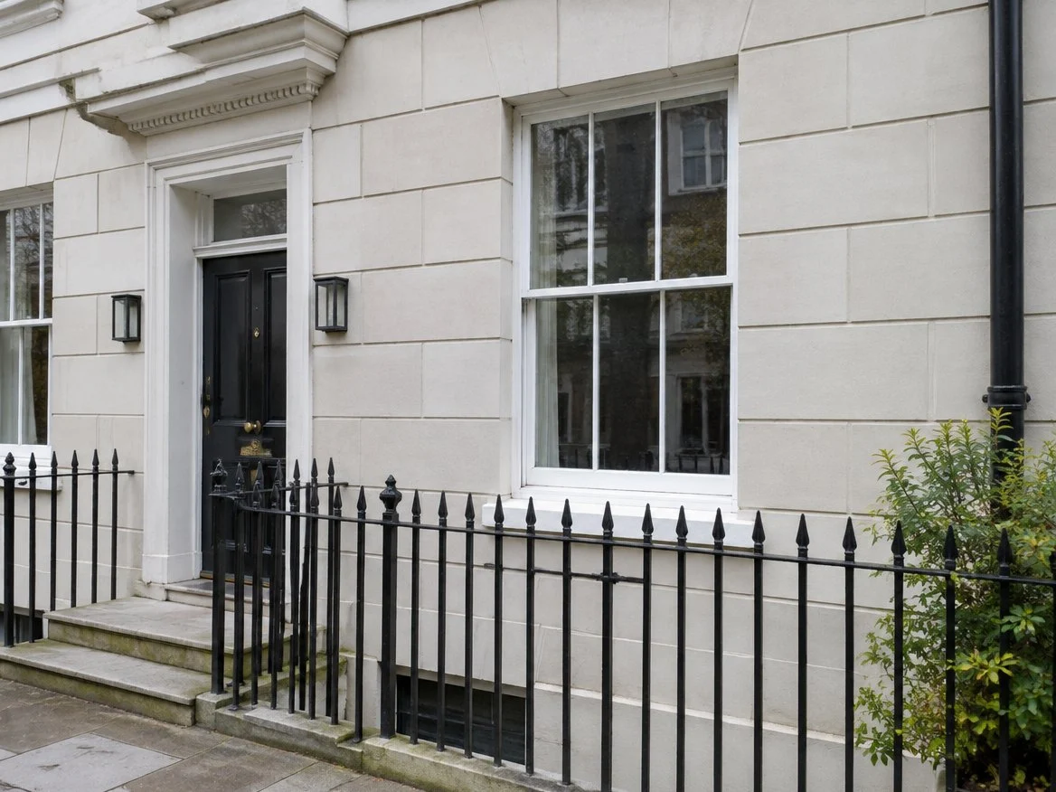 Ground floor of a terraced townhouse in pale render scored into stone blocks, with a black door, sash window and railings (illustrative image)