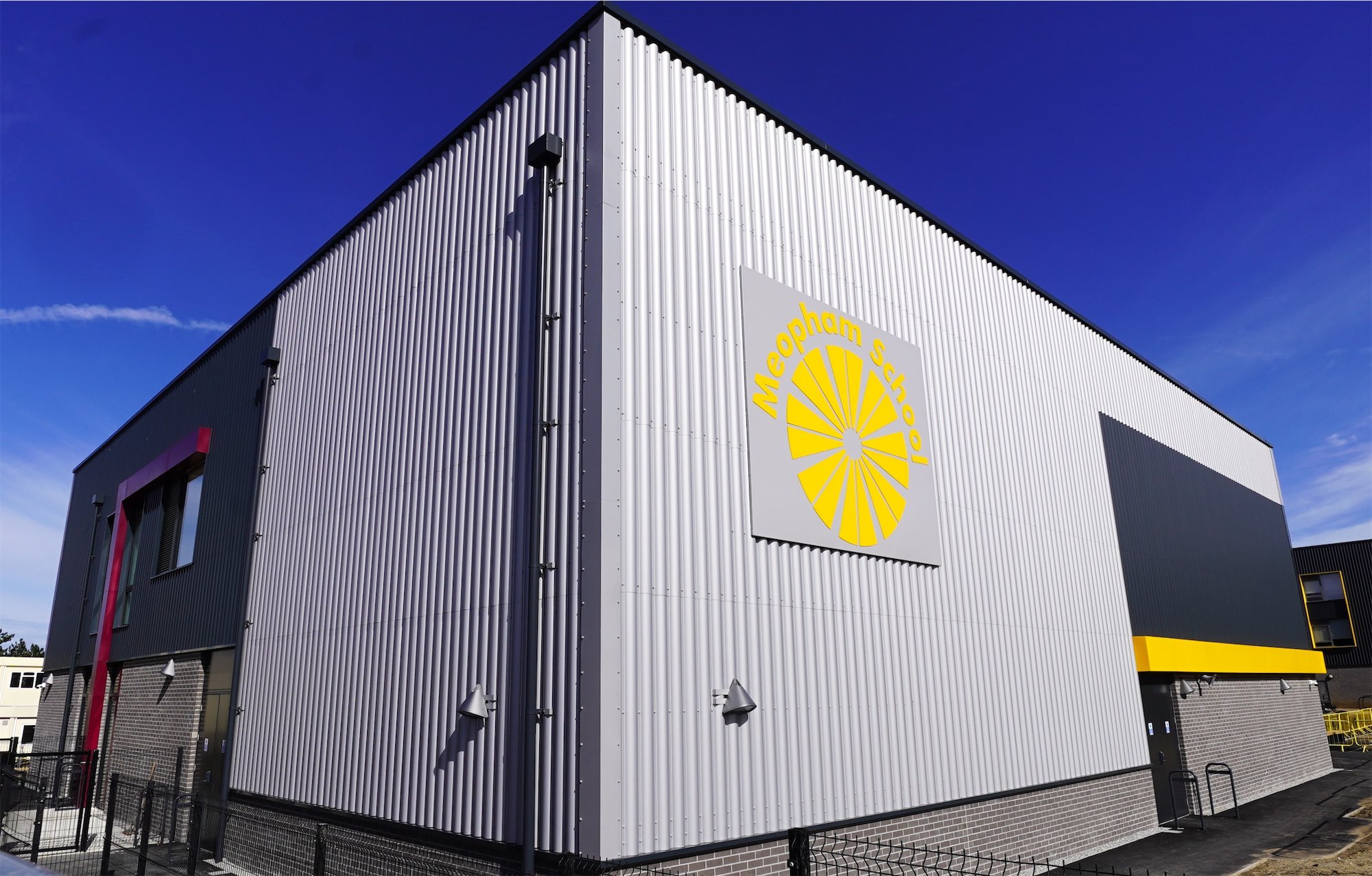 Hall block in silver profiled metal cladding with the school's sun logo, under a blue sky